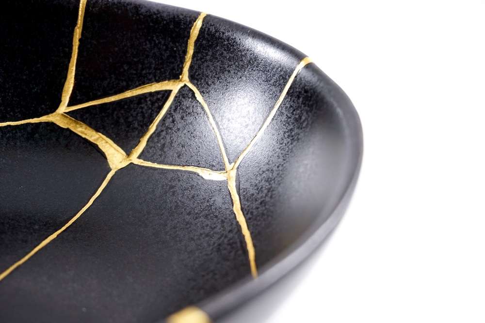 Close-up of a black ceramic bowl repaired with gold-filled cracks using the Japanese kintsugi technique against a white background.