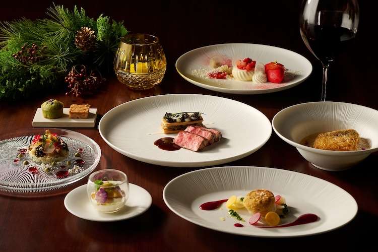A festive Christmas holiday dinner set up with wagyu beef on a white plate and another plate with sautéed scallops and a dessert in the back. A wine glass sits in the corner while a golden candle holder stands next to a Christmas tree branch.