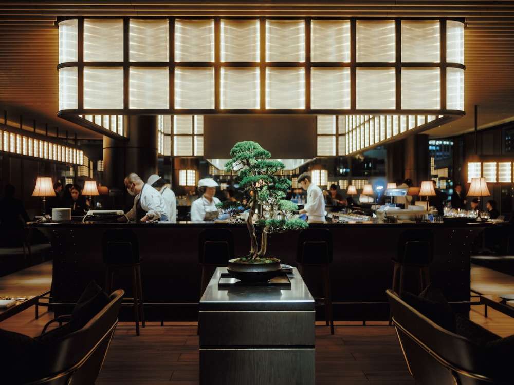 The chef grilling in an open kitchen at restaurant Janu Grill in Tokyo, Japan. The kitchen is lit with warm light and the bonsai is placed in the center.