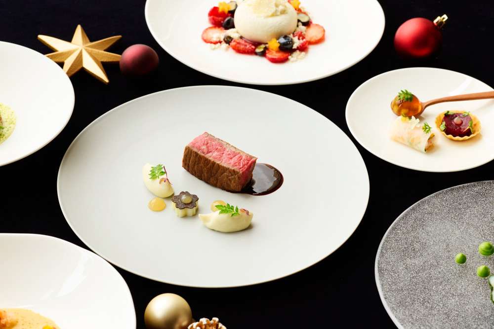 The Peninsula Hotel Tokyo's Lobby Christmas dinner course with sliced medium rare wagyu and a side of steamed white fish, and a pinecone-shaped dessert on white ceramic plates surrounded by gold ornaments.