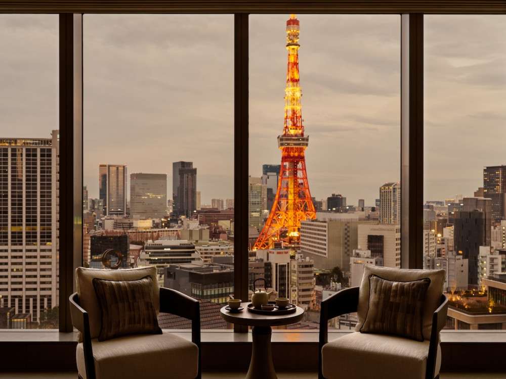A photo of a suite room of Janu Tokyo. A large window panels behind chairs show Tokyo Tower in the twilight, with orange glow.