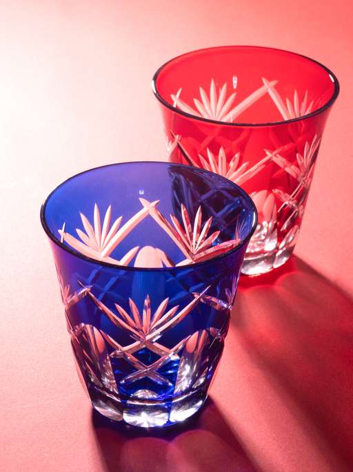 Blue and red Edo Kiriko cut glass tumblers reflect on a reddish surface, highlighting their intricate geometric patterns.