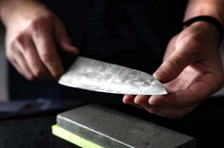 A Japanese craftsman holds a knife after sharpening its blade. Against a dark background, the silver knife glimmers in the studio. 